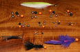 An assortment of Hareline Insta Jigs 2.0 keeled tungsten fly-tying weights in copper, black, nickel, and fluorescent orange scattered on a wood grain background alongside finished tactical jig flies.