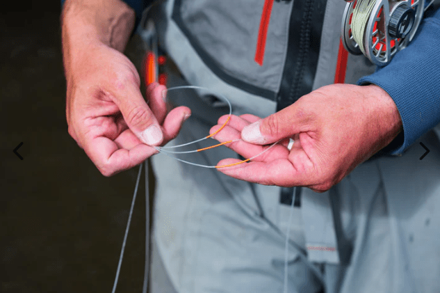 Close-up of a person's hands tying a fishing line with orange accents, wearing a gray and blue fly fishing jacket with a spool visible on the shoulder.