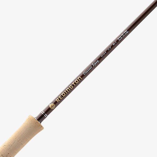 A close-up of a Redington Classic Spy fly fishing rod blank, showing the dark brown graphite shaft with gold and white branding text, and the natural cork handle on a light gray background.