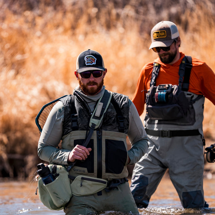 Two men in fly-fishing gear wading through shallow water, with dry reeds in the background. The man in front wears a gray and black chest rig with a water bottle pouch, while the man behind wears an orange shirt and gray overalls.