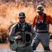 Two men in fly-fishing gear wading through shallow water, with dry reeds in the background. The man in front wears a gray and black chest rig with a water bottle pouch, while the man behind wears an orange shirt and gray overalls.