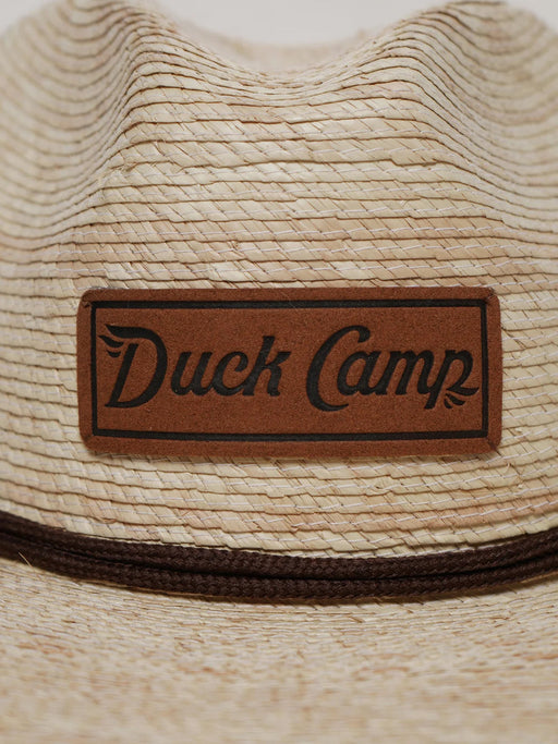 Close-up detail shot of the Duck Camp Crushable Flats Hat, focusing on the textured, tightly woven natural palm fibers and the rectangular genuine brown leather patch stamped with the "Duck Camp Premium Outdoor Goods" logo.