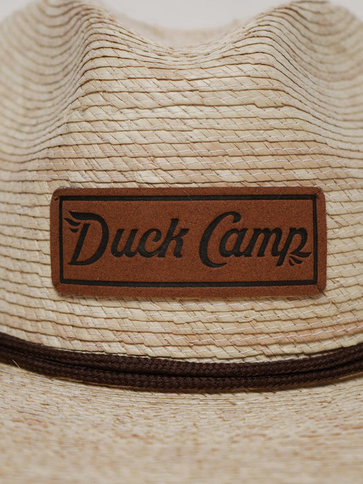 Close-up detail shot of the Duck Camp Crushable Flats Hat, focusing on the textured, tightly woven natural palm fibers and the rectangular genuine brown leather patch stamped with the "Duck Camp Premium Outdoor Goods" logo.