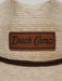 Close-up detail shot of the Duck Camp Crushable Flats Hat, focusing on the textured, tightly woven natural palm fibers and the rectangular genuine brown leather patch stamped with the "Duck Camp Premium Outdoor Goods" logo.