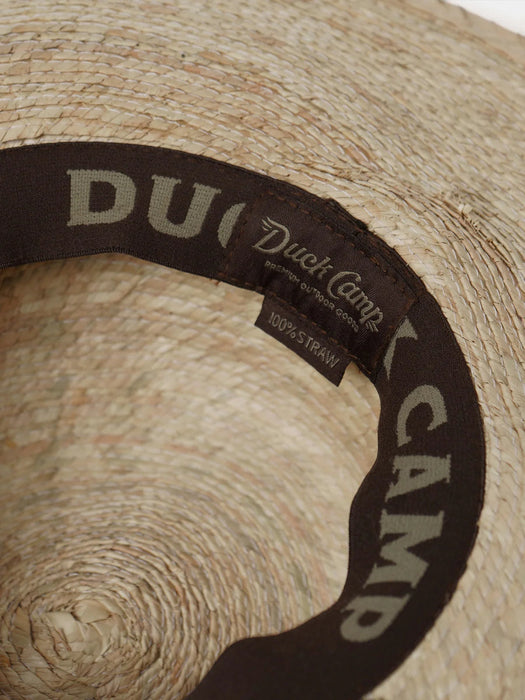 Interior view of the Duck Camp Crushable Flats Hat, highlighting the comfortable dark brown moisture-wicking sweatband printed with the repeating "DUCK CAMP" brand name and a "100% STRAW" fabric tag.