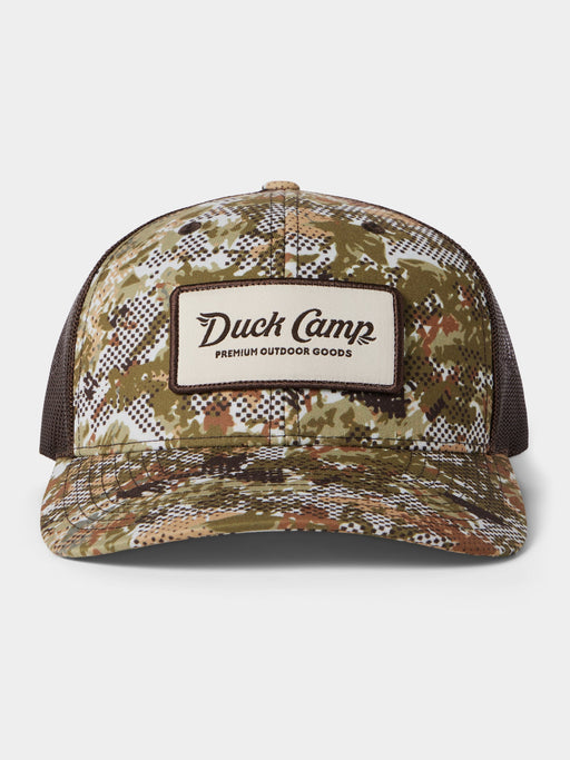 Front view of the Duck Camp Trucker Hat in Midland camo pattern, highlighting the structured front panels, the retro brown and white Duck Camp Premium Outdoor Goods logo patch, and the matching dark brown mesh back.