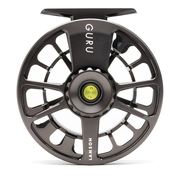 A close-up studio shot of a fly fishing reel spool, featuring a dark metallic gray body with cutout spokes, the brand name 'GURU' printed vertically on the top arm, and 'LAMSON' on the lower arm, with a small yellow-green center button.