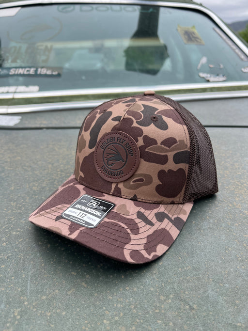 Front profile view of the Golden Fly Shop Hat 112 Bark Duck Camo resting on a car hood showcasing the structured camouflage crown and circular stitched leather fly logo patch