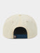 Back view of a cream-colored snapback cap with a dark blue adjustable strap and a small silver metal D-ring closure, shown on a light gray background.