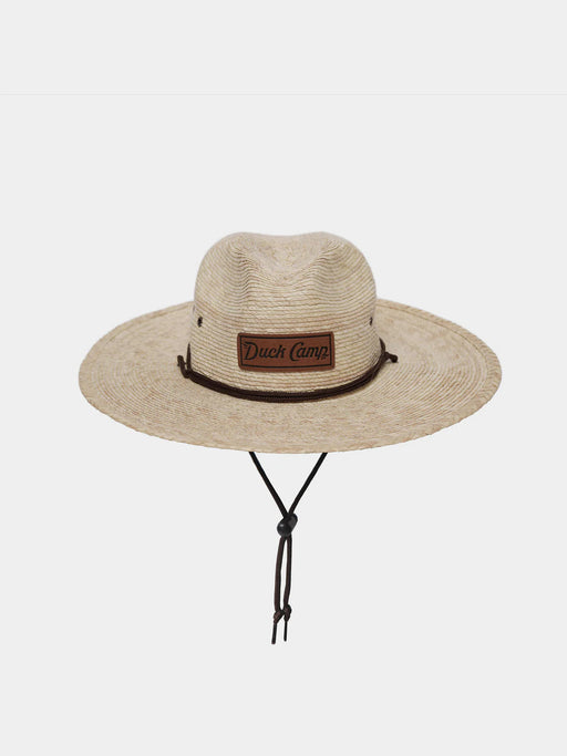 Front studio shot of the Duck Camp Crushable Flats Hat, showcasing its wide, protective woven palm straw brim, tan adjustable chin strap with toggle, and the genuine leather Duck Camp logo patch on the front crown.