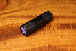 A compact Hareline UV Cure Light flashlight with a knurled black aluminum body, emitting a purple ultraviolet light beam onto a natural wood grain fly tying bench.
