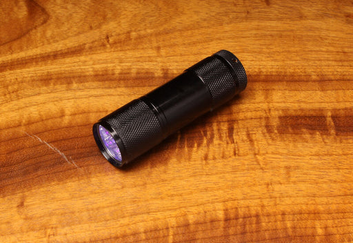 A compact Hareline UV Cure Light flashlight with a knurled black aluminum body, emitting a purple ultraviolet light beam onto a natural wood grain fly tying bench.