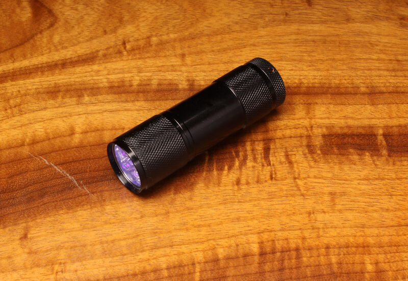 A compact Hareline UV Cure Light flashlight with a knurled black aluminum body, emitting a purple ultraviolet light beam onto a natural wood grain fly tying bench.