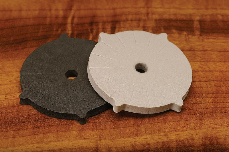 The Hareline Vise Fly Racks 2-Pack displaying one charcoal black and one off-white circular foam rack wheel with 16 perimeter slots resting on a wooden fly tying bench.